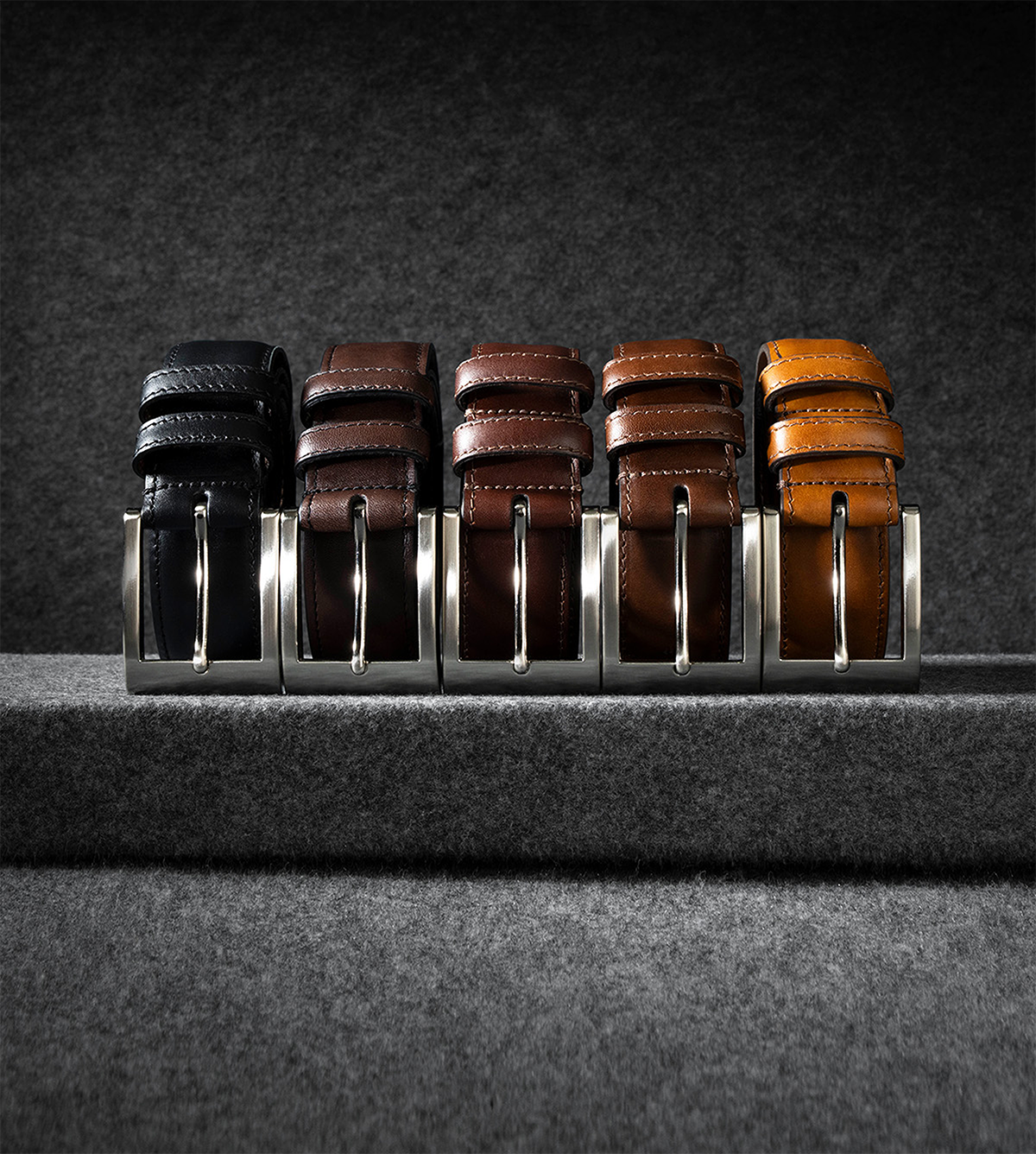 Five men’s leather belts displayed next to each other. One is black and the others are various shades of brown. Cinq ceintures en cuir pour hommes exposées les unes à côté des autres. L'un est noir et l'autre