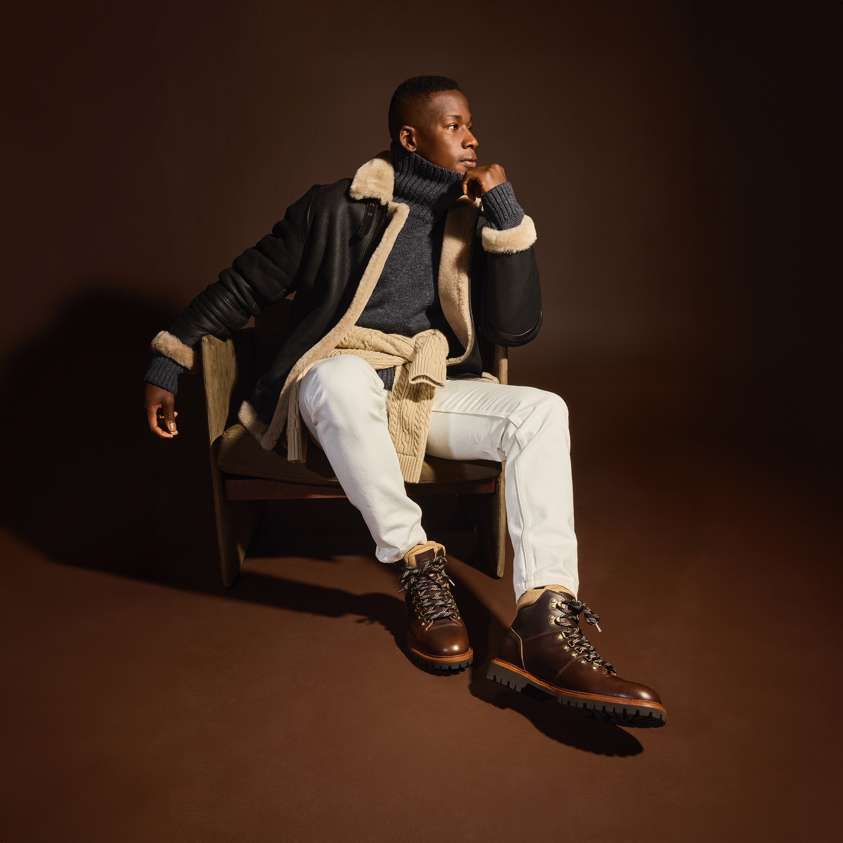 A man sitting in a chair modeling off-white jeans, a gray turtleneck sweater, a black bomber jacket, and a pair of brown leather boots. Un homme assis sur une chaise modélisant un jean blanc cassé, un pull à col roulé gris, une veste bomber noire et une paire de bottes en cuir marron.