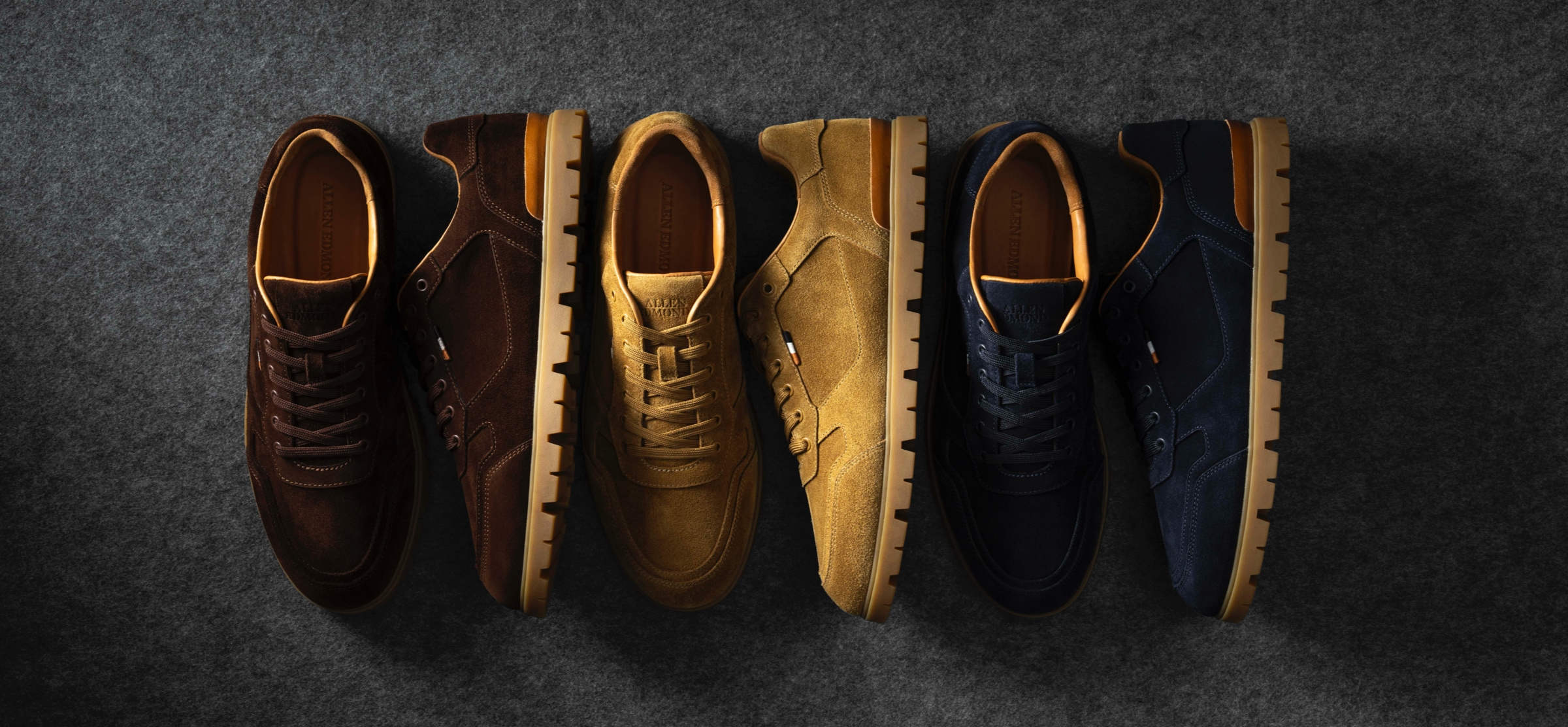 A top view of three pairs of men’s suede sneakers in the colors brown, tan, and navy blue. / Vue de dessus de trois paires de baskets en daim pour hommes, dans les couleurs marron, beige et bleu marine.