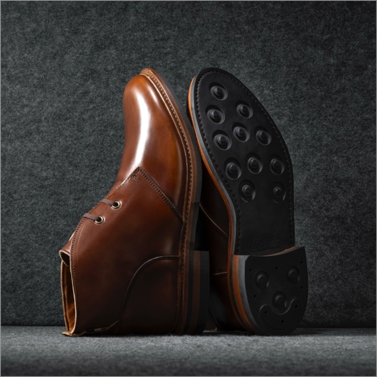 A pair of men’s brown dress boots displayed vertically with a view of the Dainite rubber sole. / Une paire de bottines habillées marron pour hommes présentée verticalement avec une vue de la semelle en caoutchouc Dainite.