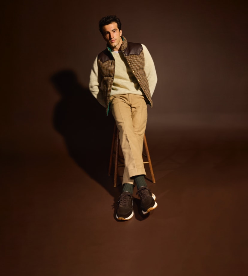 A man leaning against a stool modeling a pair of brown Allen Edmonds sneakers. / Un homme appuyé contre un tabouret portant une paire de baskets marron Allen Edmonds.