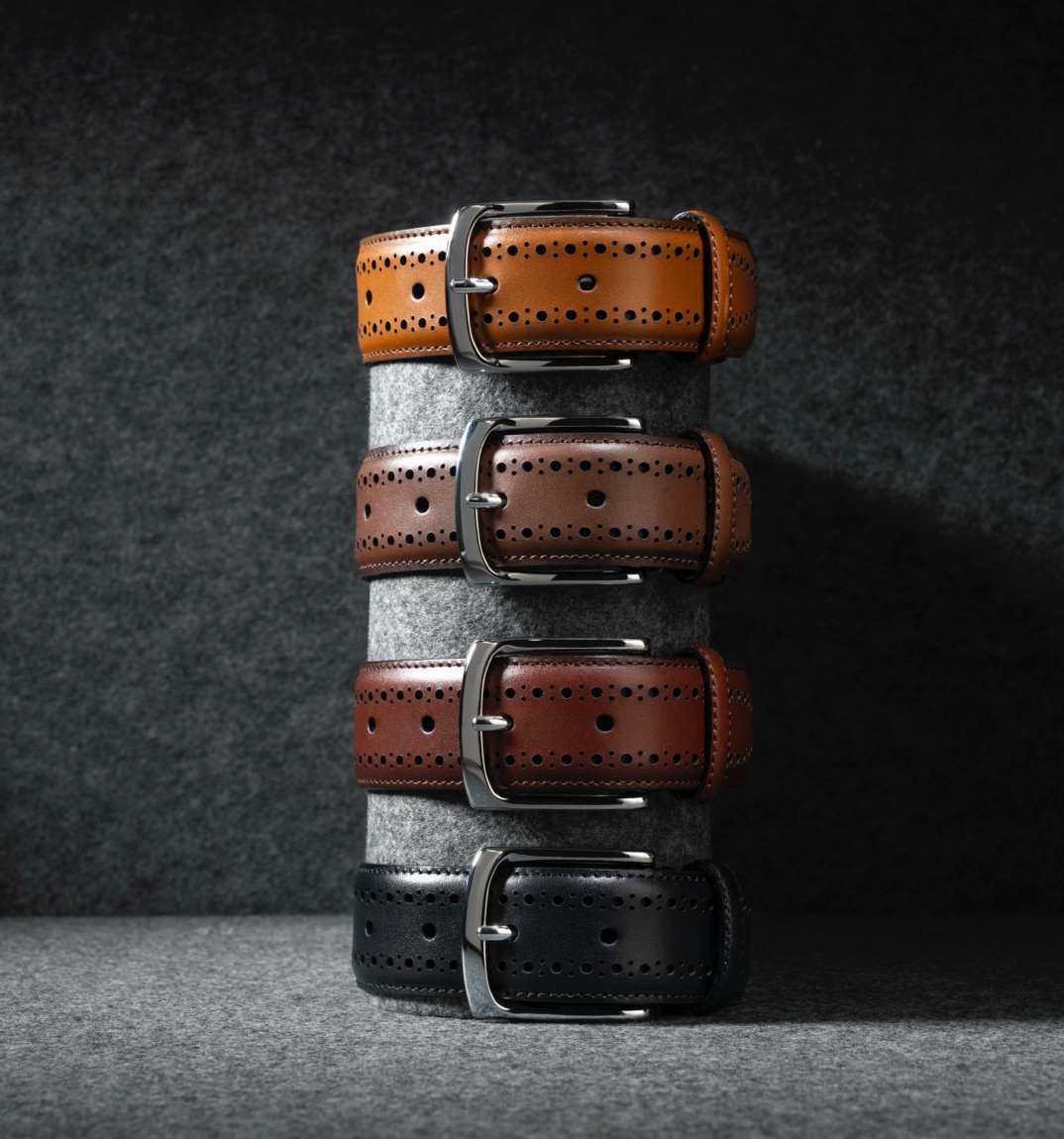 Four men’s leather belts stacked on top of one another in black and various shades of brown.  Quatre ceintures en cuir pour hommes empilées les unes sur les autres, de couleur noire et de différentes nuances de marron.