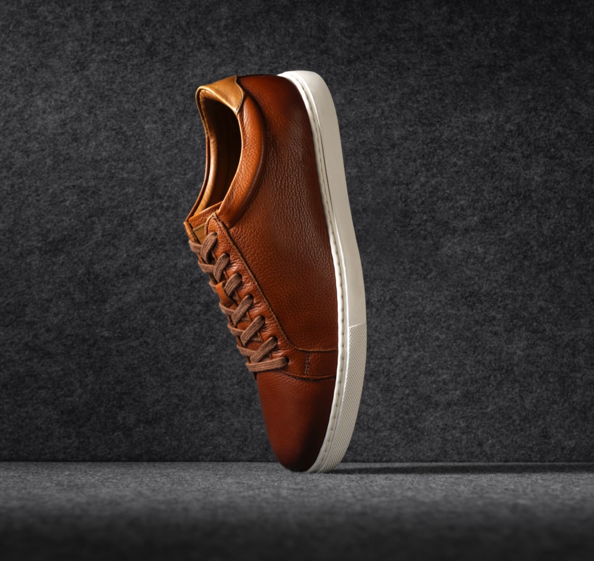 A men’s brown leather sneaker displayed vertically on its toe. Une sneaker en cuir brun pour homme présentée verticalement sur son bout.