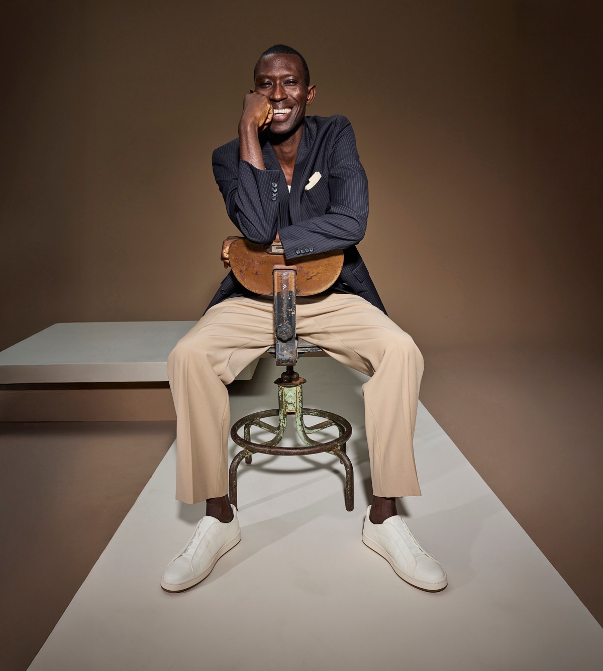 Man seated on a vintage stool and wearing white slip-on sneakers with beige trousers and a navy pinstripe suit jacket 